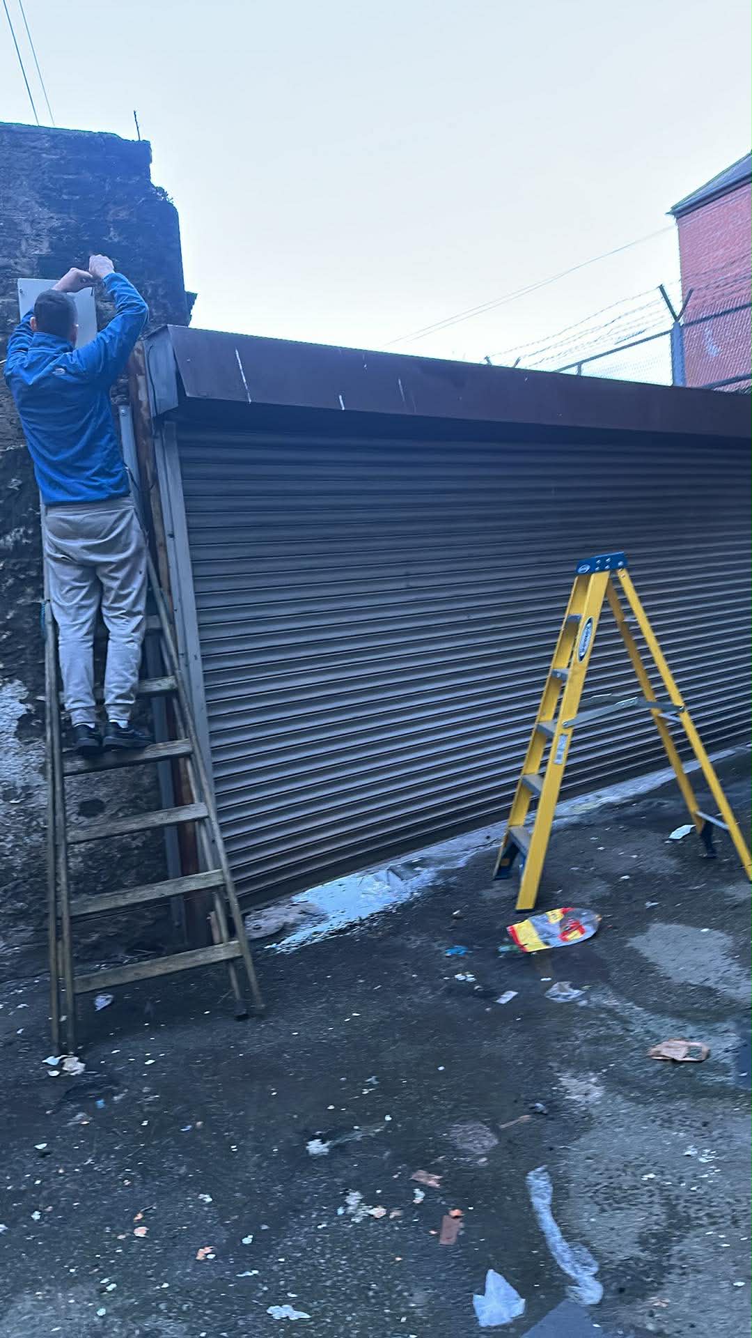 SteelMotion engineer on a ladder repairing a large dark commercial roller shutter in a Dublin laneway at dusk