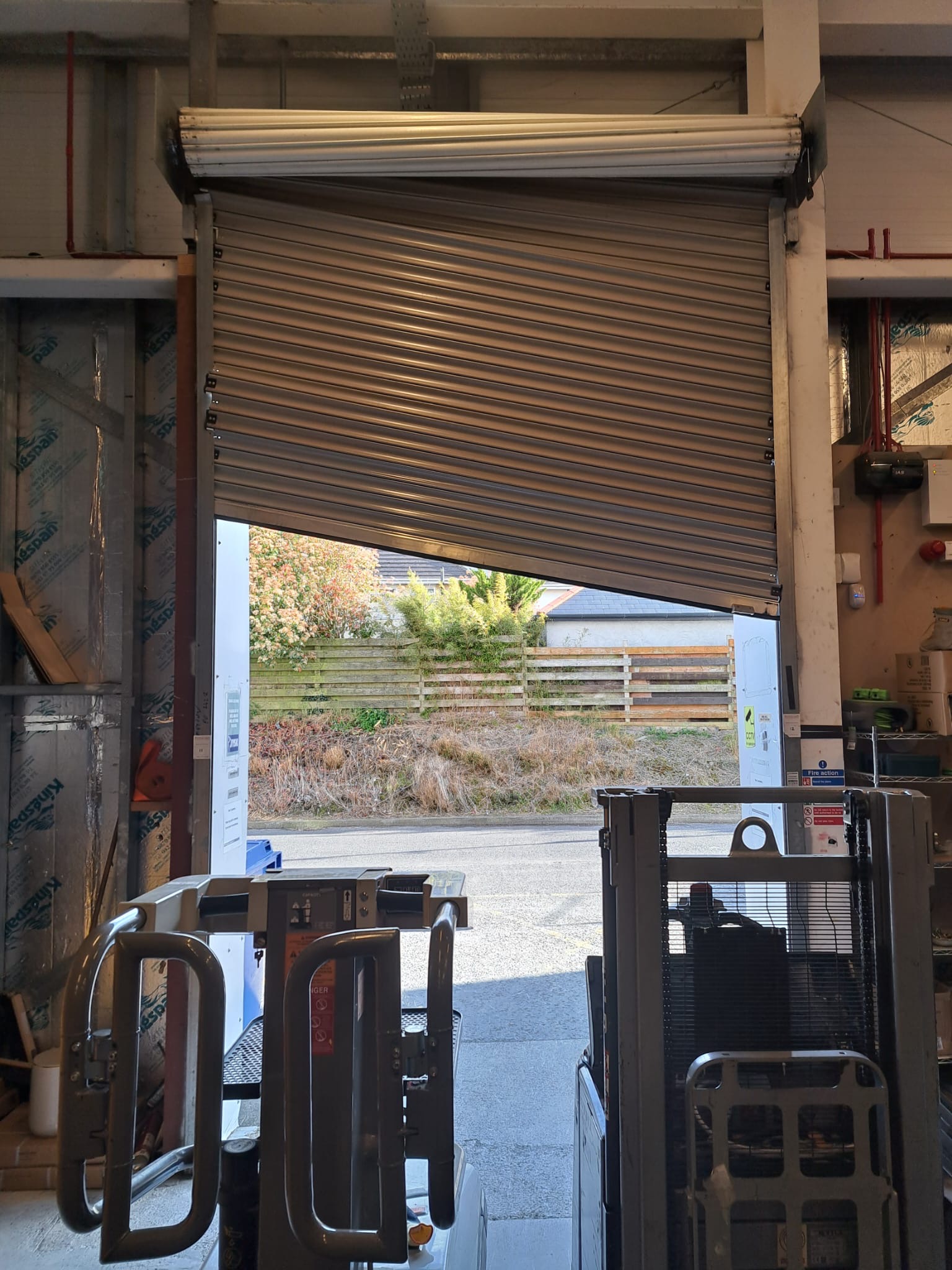 Beige industrial roller shutter loading-bay door part-raised inside a Dublin warehouse unit, with forklifts in the foreground